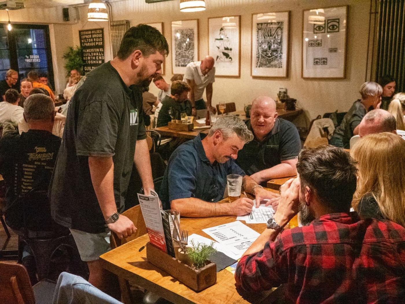Teams filling in answer sheets during the Big Business Pub Quiz at Croxtons, with staff talking to a table