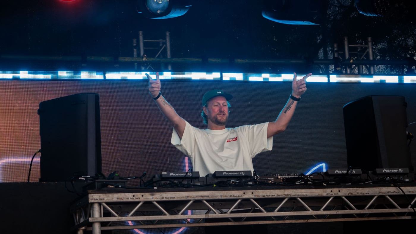 A DJ on the Send It 100 main stage with both arms raised, lit blue against the LED wall